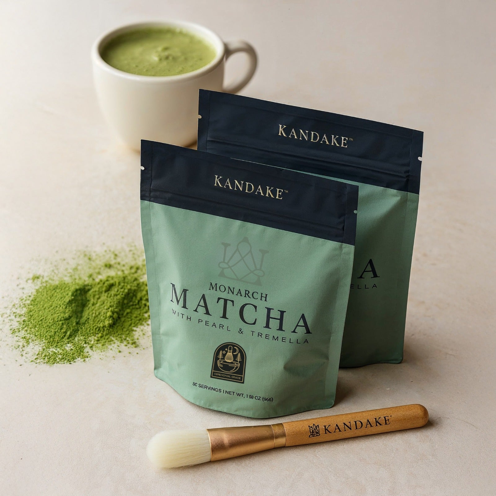 Two green Kandake matcha packages with a cup of matcha and the kandake monarch matcha brush on a light background